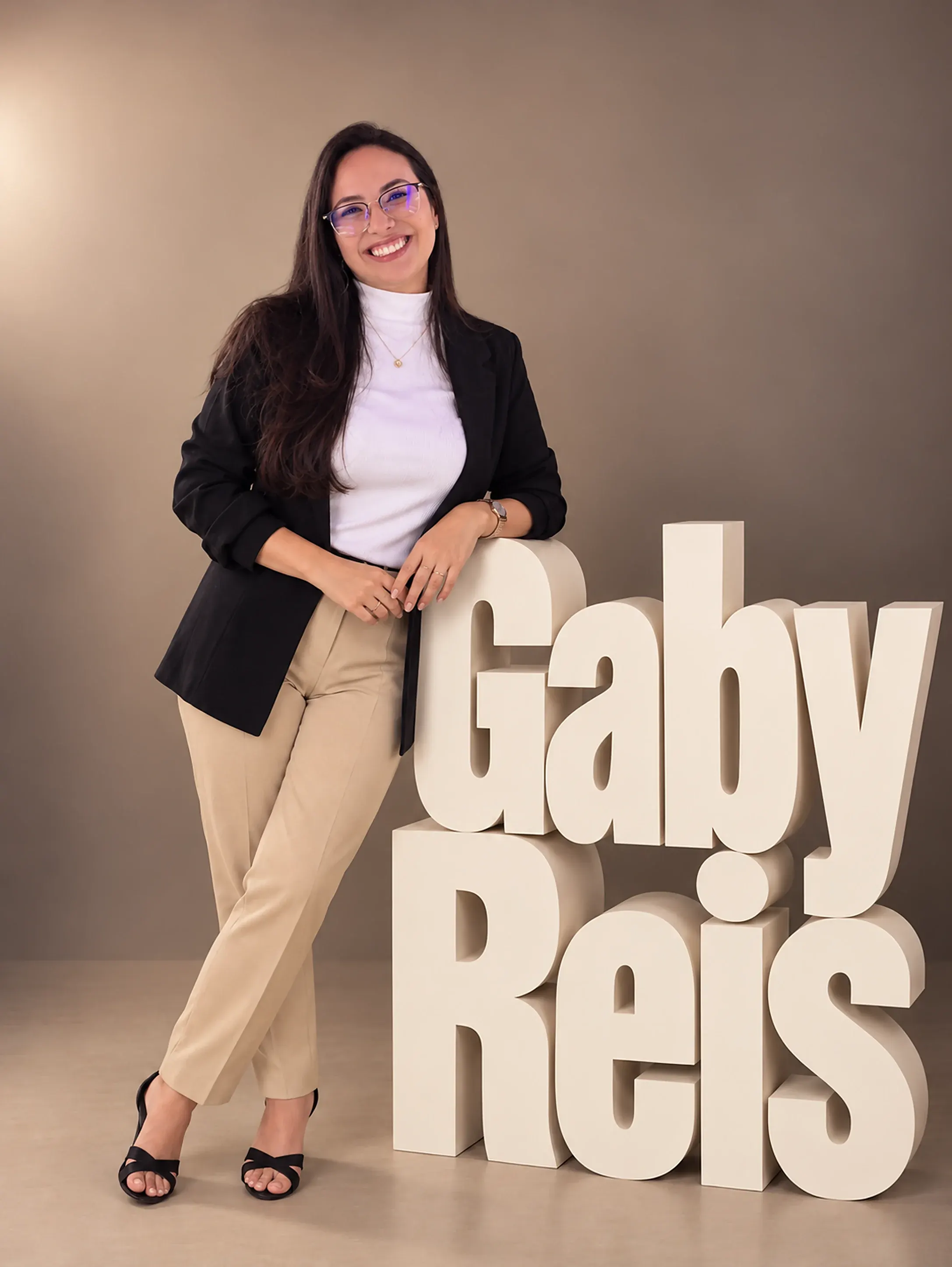 Gabrielly Reis sorrindo, de blazer preto, apoiada nas letras do nome Gaby Reis